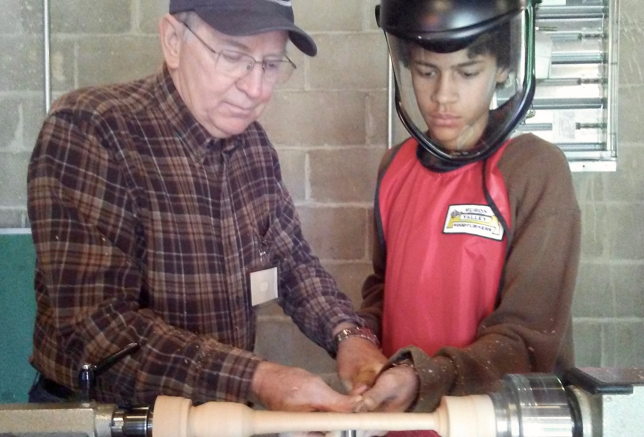 an older man in a wood shop showing a teenage boy, who is wearing a face shield, how to use a lathe