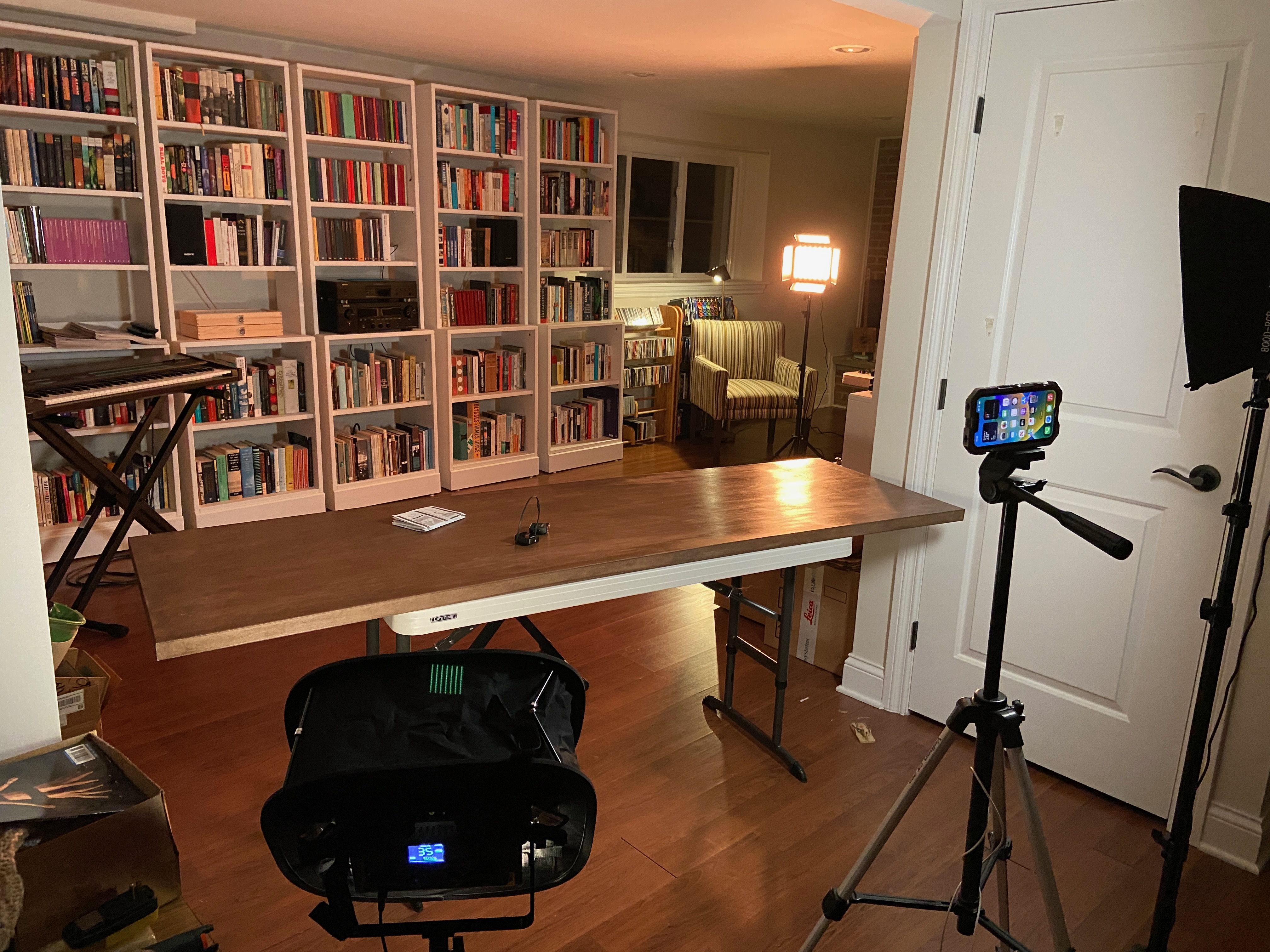 a view of the basement library shot in the opposite direction, showing a table, an iPhone on a tripod, lighting gear, and boxes